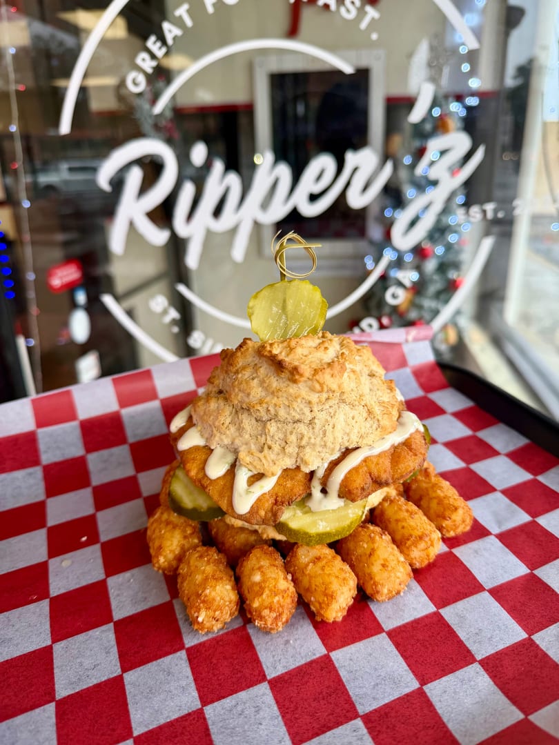 A fried chicken biscuit sandwich with pickles and sauce, surrounded by tater tots on checkered paper.