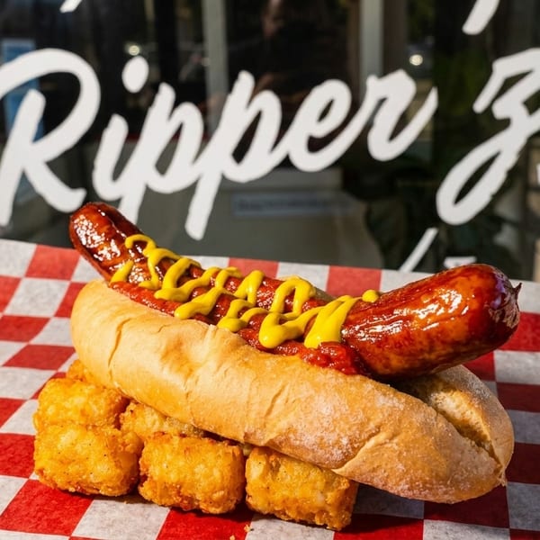 A hot dog with mustard and relish on a bun with tater tots, in front of a window with "Ripper Z" written on it.