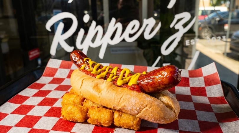 A hot dog with mustard and relish on a bun with tater tots, in front of a window with "Ripper Z" written on it.