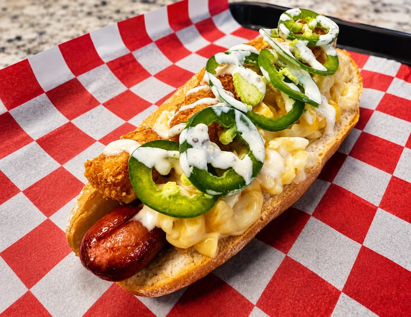 Hot dog in a bun with mac and cheese, fried chicken, jalapeños, and white sauce.