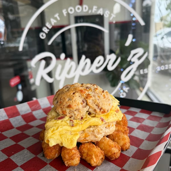 A breakfast sandwich with egg and biscuit, surrounded by tater tots on red and white checkered paper.