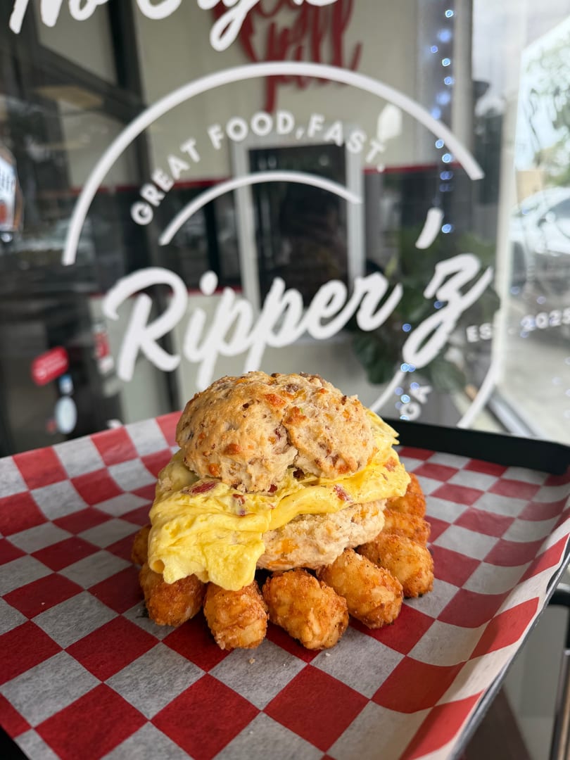A breakfast sandwich with egg and biscuit, surrounded by tater tots on red and white checkered paper.