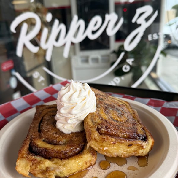 Two cinnamon rolls with whipped cream and syrup on a plate, with a restaurant window in the background.