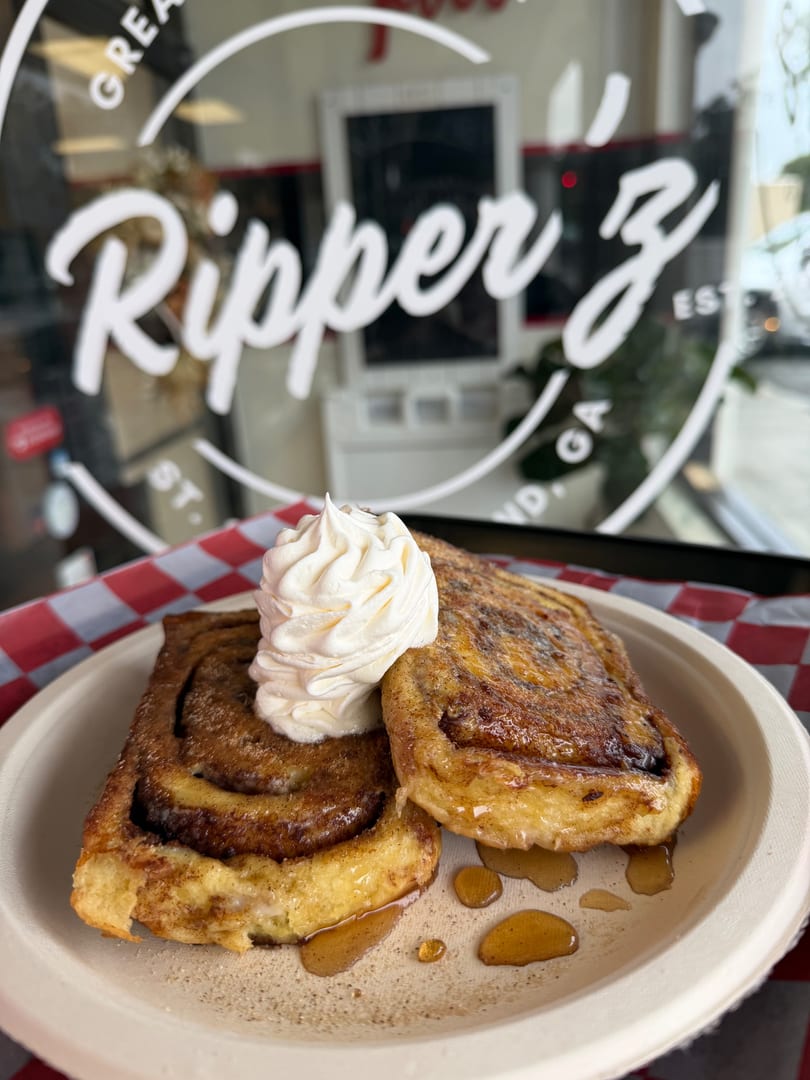 Two cinnamon rolls with whipped cream and syrup on a plate, with a restaurant window in the background.